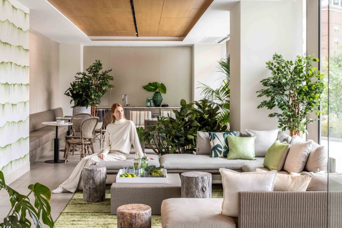 Bright contemporary lounge with a neutral sectional sofa, natural wood ceiling, abundant greenery, and large windows. A person sits near a coffee table surrounded by plants, textured furnishings, and earthy wood accents.