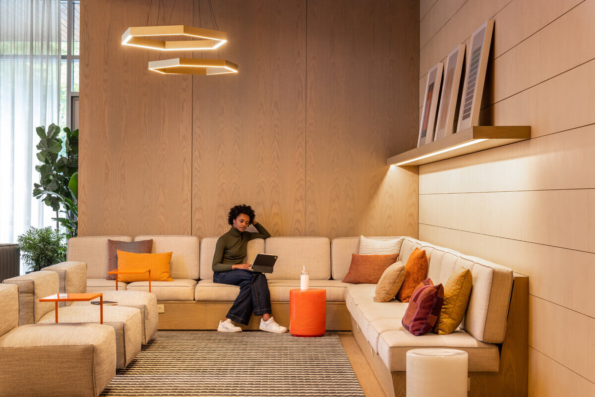 Contemporary lobby seating area with a warm wood-paneled interior, cream sectional sofa, colorful accent pillows, and orange side tables. A person sits on the sofa using a tablet, while geometric pendant lights and built-in shelving add visual interest.