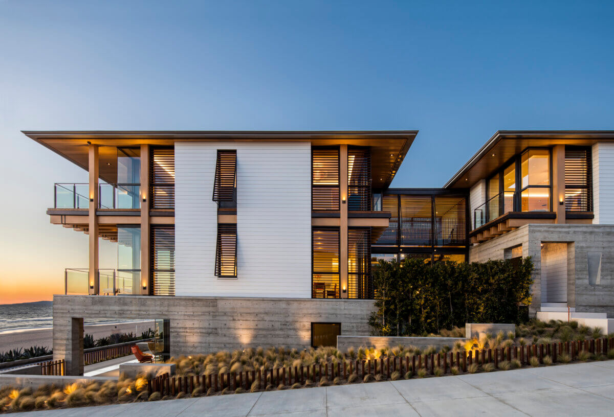 Modern oceanfront residence featuring white exterior cladding, concrete walls, expansive windows, and wood accents. Illuminated interiors and landscaped grasses create a warm contrast against the blue dusk sky and ocean backdrop.
