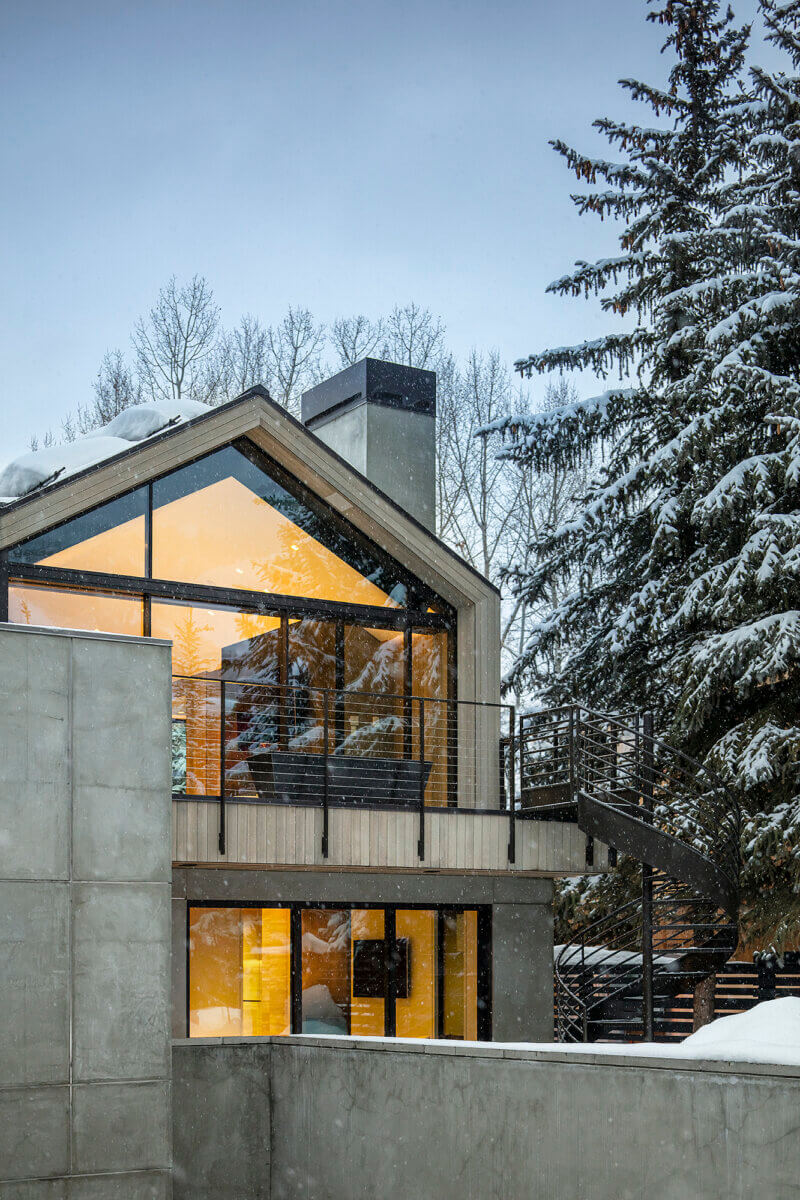 Contemporary mountain home with a steeply pitched roof, expansive triangular glazing, concrete walls, and a black metal exterior staircase. Warm interior lighting contrasts with the snow-covered roof and surrounding evergreen trees.