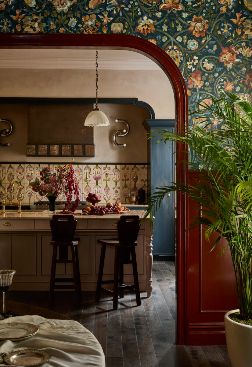 Eclectic kitchen and dining interior framed by a deep red arched doorway, with floral wallpaper, patterned tile backsplash, dark wood floors, and blue cabinetry. A large potted palm, decorative lighting, and richly colored finishes add a layered, traditional character.