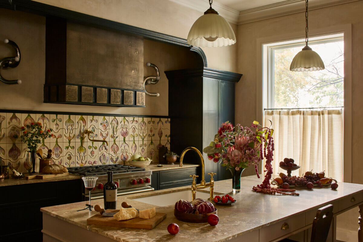 Warm, traditional kitchen with a large stone island, dark cabinetry, brass fixtures, and an ornate patterned tile backsplash. Floral arrangements and fruit decorate the countertops, while two scalloped pendant lights and a large window add warmth and natural light.