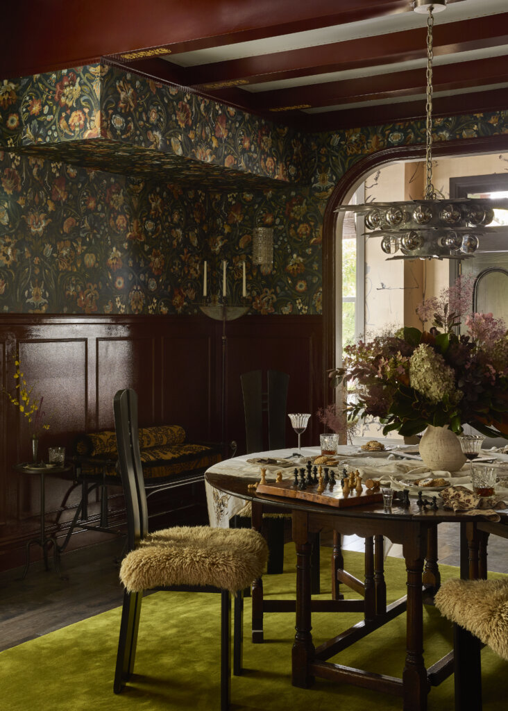 Eclectic, moody dining room with a dark wooden gateleg table set with a chessboard, pastries, crystal glasses of red wine, and a large arrangement of hydrangeas and smoke bush, flanked by two high-backed Mackintosh-style chairs with shaggy ochre sheepskin seats on a moss-green rug. Dark mahogany wainscoting and navy William Morris-style floral wallpaper line the walls beneath a chrome sphere chandelier, and a curved archway opens to a brighter chinoiserie-style room.