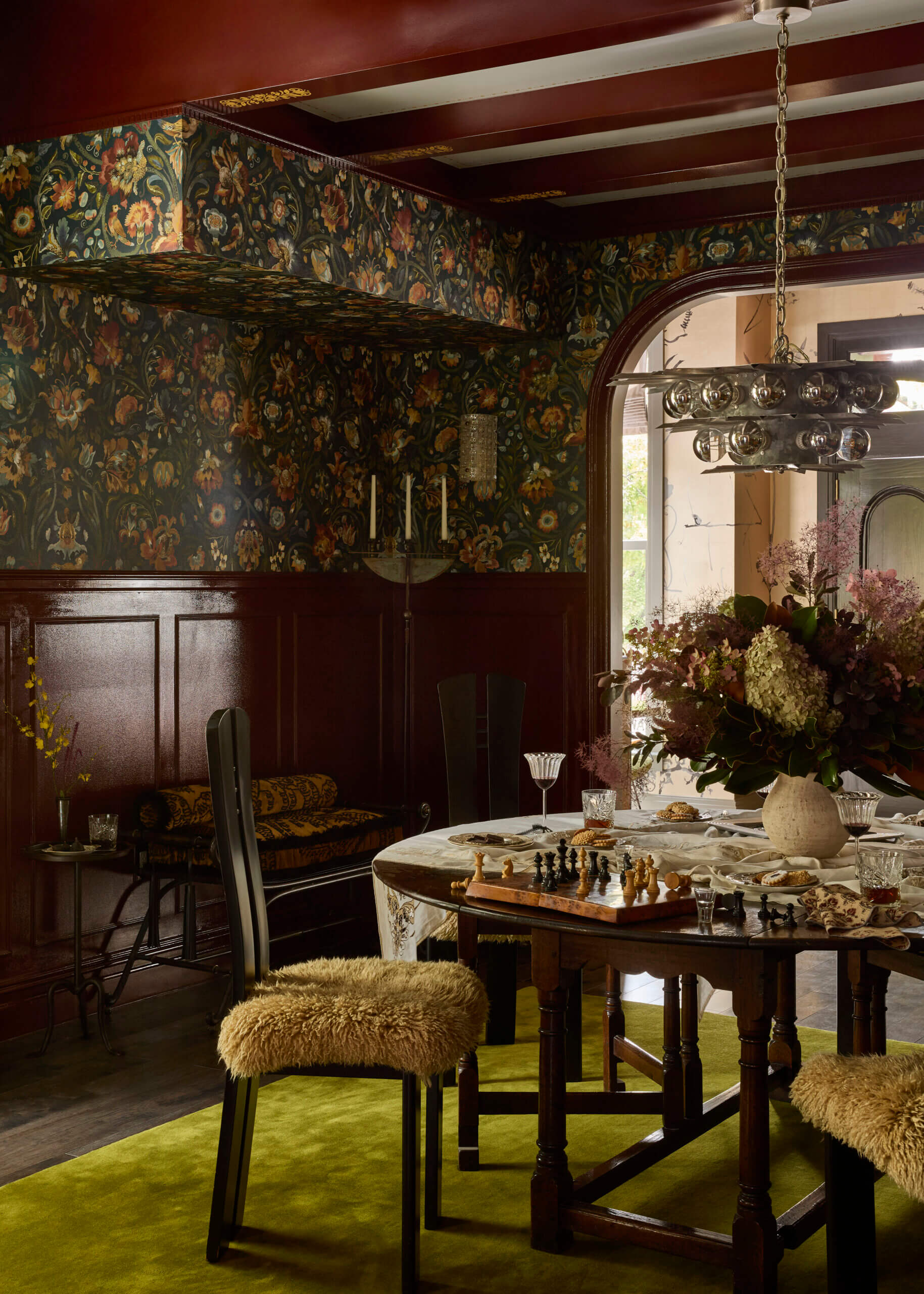 Eclectic, moody dining room with a dark wooden gateleg table set with a chessboard, pastries, crystal glasses of red wine, and a large arrangement of hydrangeas and smoke bush, flanked by two high-backed Mackintosh-style chairs with shaggy ochre sheepskin seats on a moss-green rug. Dark mahogany wainscoting and navy William Morris-style floral wallpaper line the walls beneath a chrome sphere chandelier, and a curved archway opens to a brighter chinoiserie-style room.