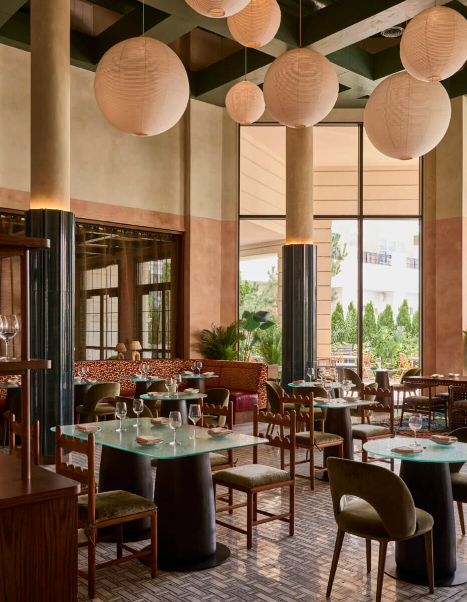 Elegant restaurant interior with globe-shaped pendant lights, tall columns, patterned banquette seating, glass dining tables, and sculptural wood chairs. Warm neutral tones, dark green ceiling beams, and large windows create a refined, inviting atmosphere.