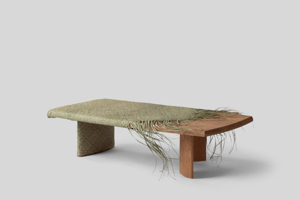 A low wooden table with a natural woven fiber surface, partially unraveling into loose strands that drape over the edges, combining traditional craftsmanship with an organic aesthetic.