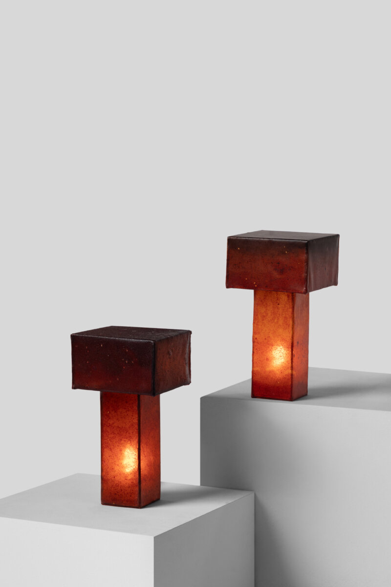 Two sculptural table lamps with deep reddish-brown, rectangular shades and slender rectangular bases. The translucent bases emit a warm amber glow, creating a soft, atmospheric light.