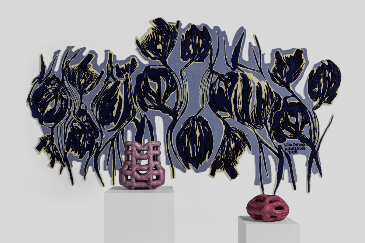 A large, intricate botanical textile wall piece featuring stylized dark blue flowers and foliage against a muted lavender background. Two small sculptural ceramic objects in shades of pink and mauve are displayed on white pedestals beneath it.