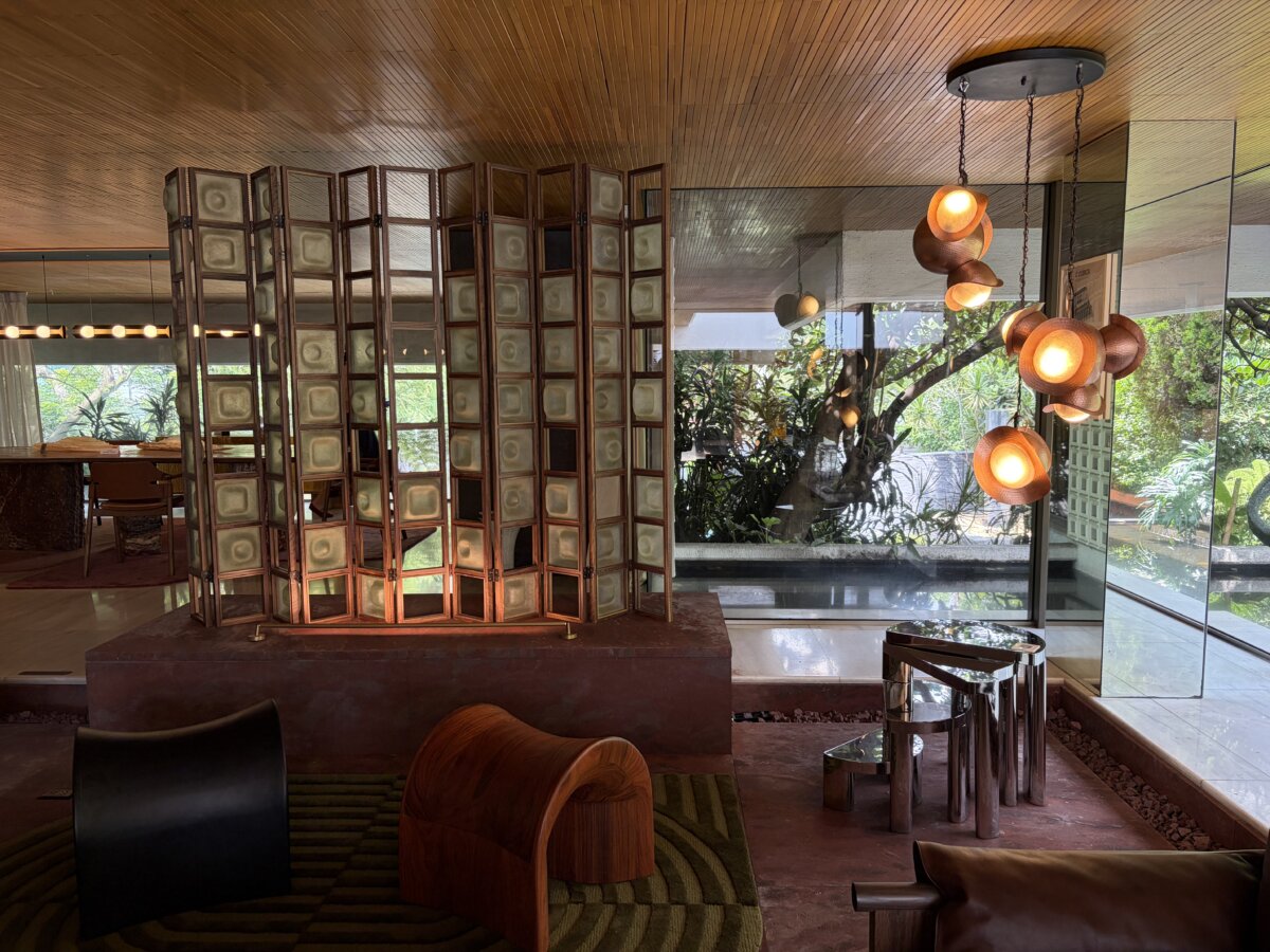 Interior of a contemporary design space featuring a sculptural folding screen with wood framing and translucent panels, surrounded by warm wood finishes and floor-to-ceiling windows overlooking lush greenery. Organic pendant lights with glowing amber shades hang above polished metal nesting tables, while sculptural lounge chairs sit on a textured area rug.