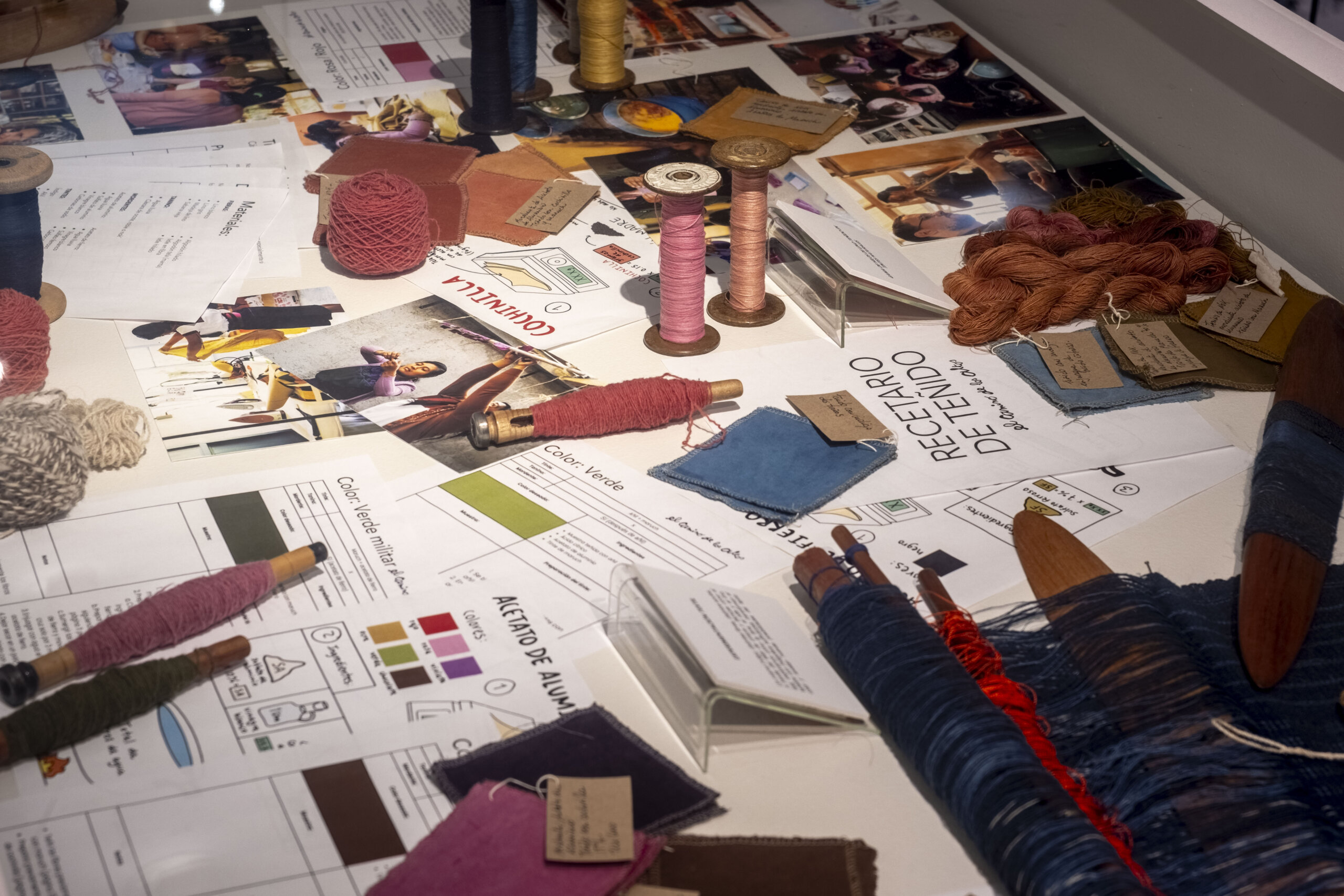 A textile design workspace displaying yarn spools, colorful fabric swatches, thread samples, photographs, and design sketches arranged across a table.