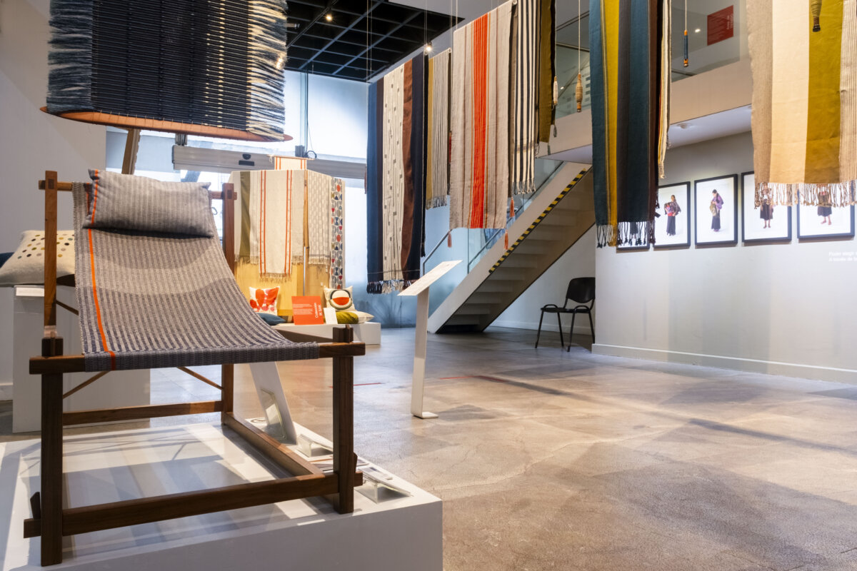extile exhibition featuring hanging fabric samples in varied colors, patterns, and textures, alongside a woven lounge chair on a wooden frame and a large suspended textile installation.