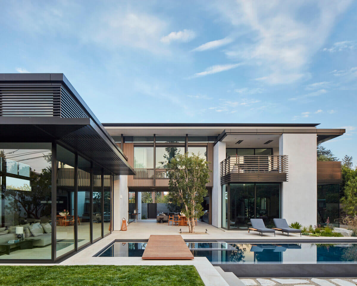 Contemporary luxury residence with clean geometric lines, white stucco walls, dark metal louvers, and expansive glass windows surrounding a swimming pool. A wood deck, landscaped garden, and outdoor lounge chairs complement the minimalist architecture.