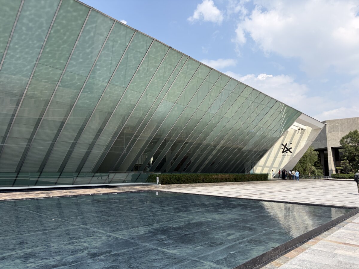 Exterior view of a contemporary museum featuring a dramatic angular glass façade with sloping geometric lines. A shallow reflecting pool extends across the foreground, mirroring the building and surrounding sky, while visitors gather near the entrance beneath a bright blue sky.