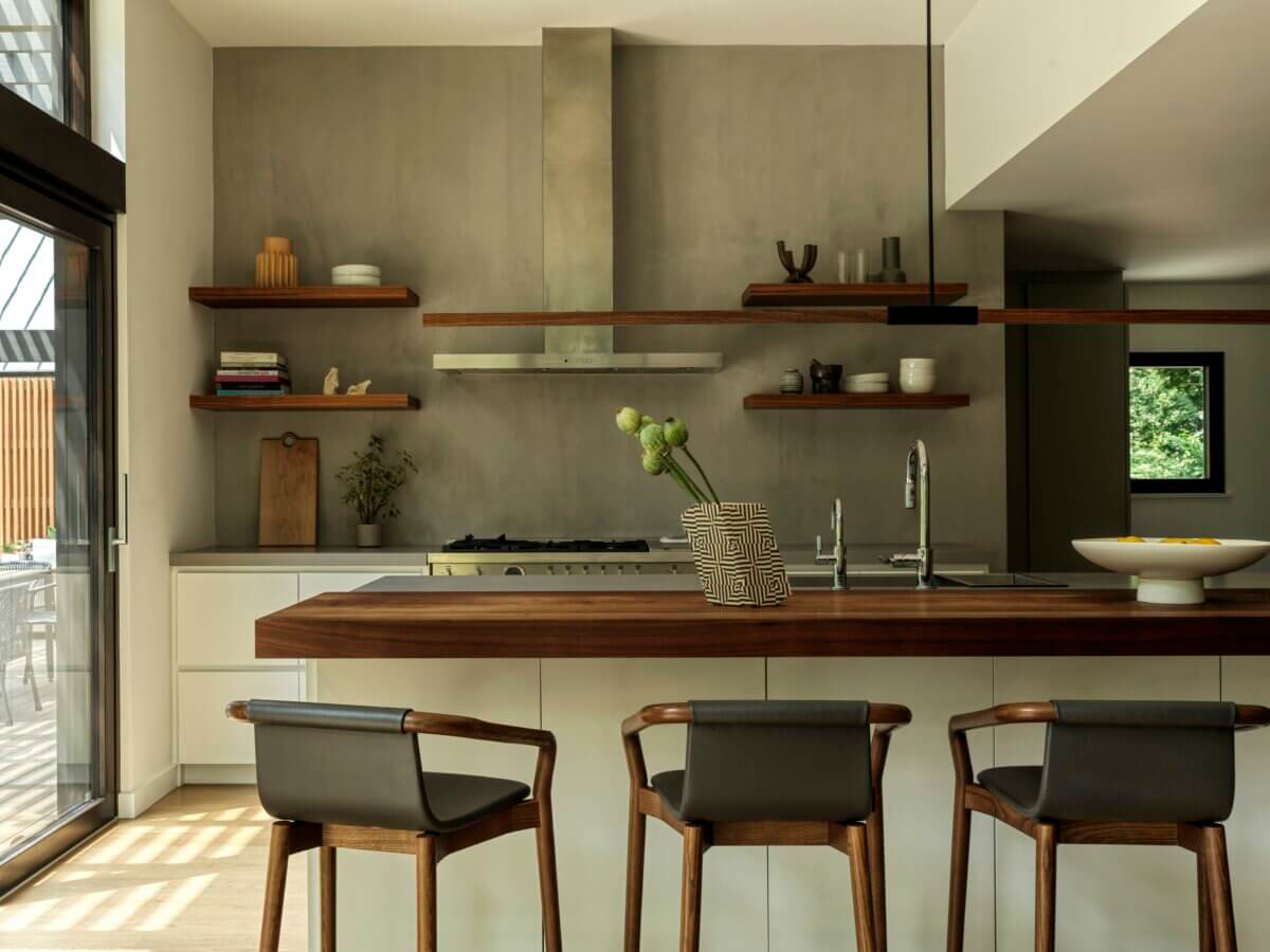 Contemporary kitchen with a warm wood island, minimalist cabinetry, open shelving, and a textured concrete wall. Three wood-and-leather counter chairs sit beneath the island, while natural light enters through large windows and glass doors.