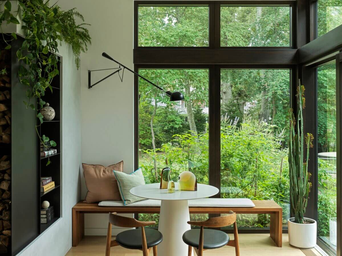 Cozy dining nook with a round white table, built-in wooden bench, sculptural glass objects, and two wood-and-black chairs. Floor-to-ceiling windows overlook lush greenery, while abundant plants and a black wall-mounted lamp add warmth and character.