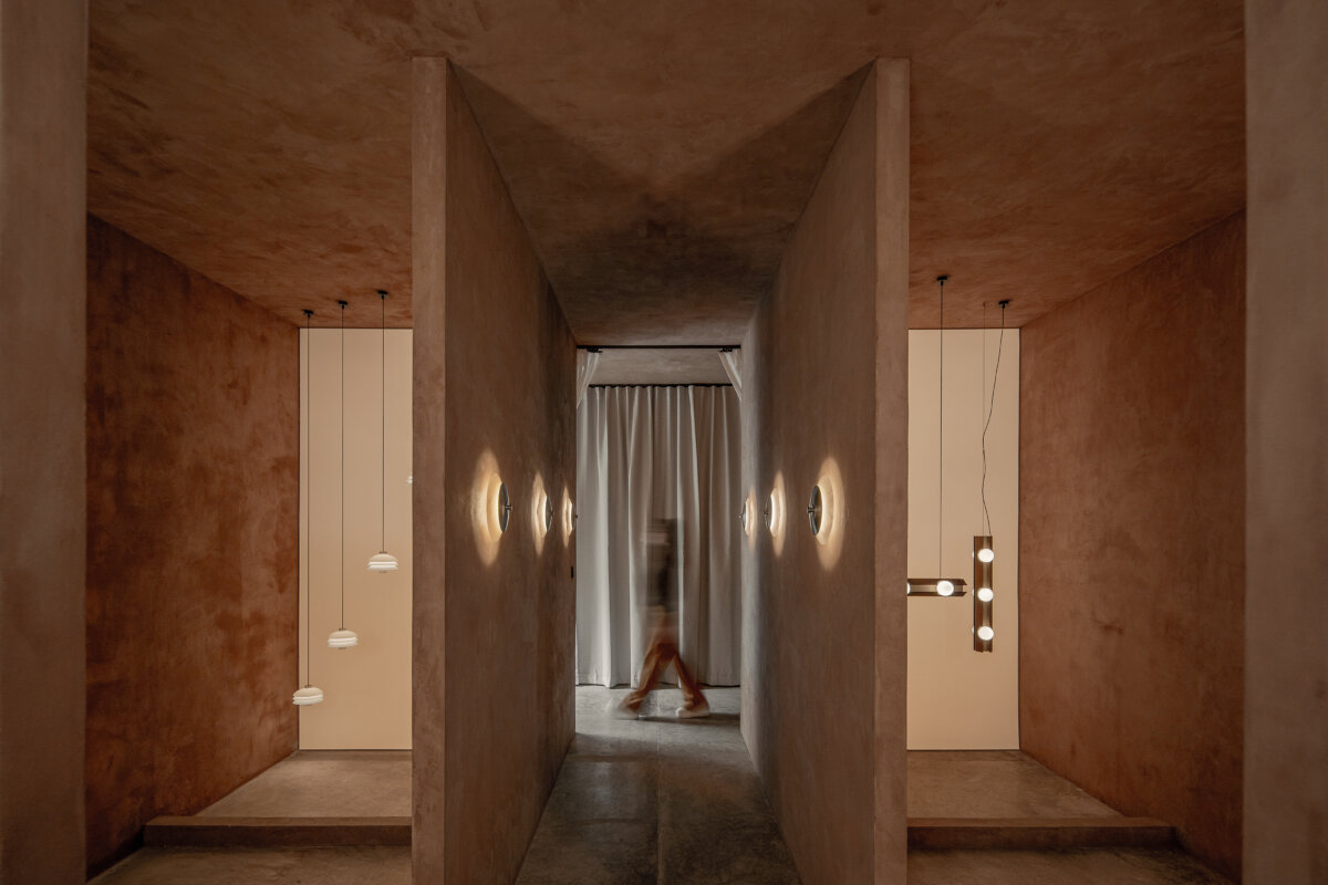 Interior of the Bandido showroom featuring sculptural pendant and wall lighting displayed within a symmetrical, earth-toned architectural space with textured terracotta walls, concrete flooring, and a central corridor.