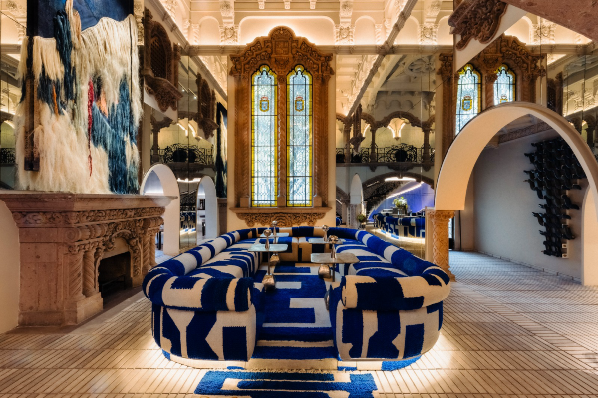 An ornate interior featuring a sculptural blue-and-cream upholstered sectional sofa with bold geometric patterns, arranged around low tables. The seating area is set within an elaborate historic space with carved wooden details, stained-glass windows, arched doorways, and a decorative stone fireplace. A large textured textile artwork hangs above the fireplace, while warm architectural lighting illuminates the tiled floor and surrounding lounge.